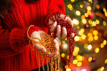 Beautiful red and golden beads close-up in hands of a young girl in warm bright red sweater. Garland light of christmas tree on the background.