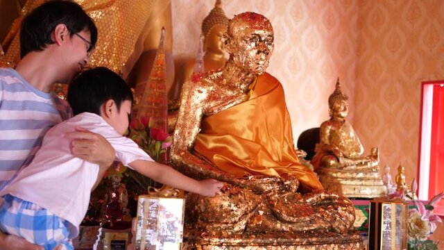 4K Buddhist asian child boy and dad apply gold leaf to buddha statue, son and father having activity together, making merit in local temple in countryside. Happy family activity in holiday.