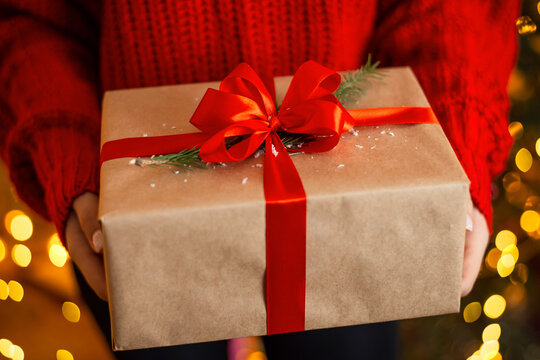 Close-up picture of big present box, wrapped in parchment paper and decorated with red ribbon and little branch of fir-tree, christmas time. Gift in hands of a girl, wearing red sweater.