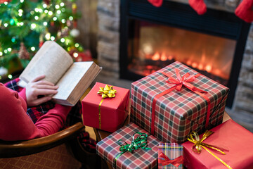 Alone woman covered plaid reading book sitting and relaxation on armchair near fareplace and christmas tree after finishing pakking gift boxes for family.