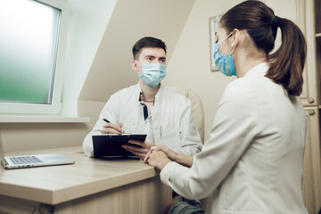 Admission to the doctor during quarantine. Young doctor and patient sitting in the office wearing protective masks. The doctor writes a prescription to a young female patient