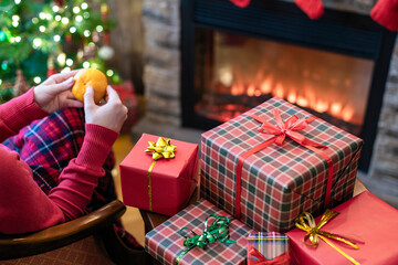 Alone woman covered plaid peels tangerine sitting and relaxation on armchair near fareplace and christmas tree after finishing pakking gift boxes for family.