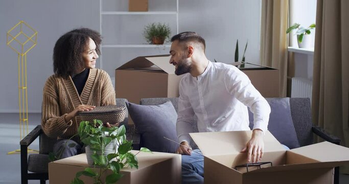 Young couple caucasian man and african american woman unpacking things in living room after moving to new home, family receives parcel by post delivery from courier open cardboard box take out dishes