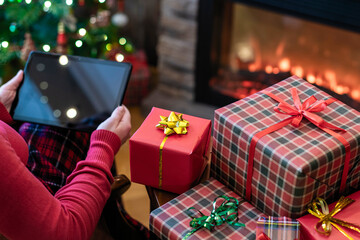 Christmas. Woman in sweater using tablet for searching gift ideas sitting at table near fireplace and christmas tree. Concept