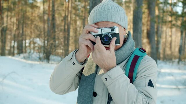 Portrait of senior man taking pictures with camera outdoors in winter forest smiling using modern device. Photography and travelling in winderness concept.