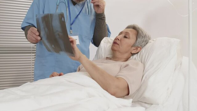 Medium shot of senior Asian woman lying in bed in hospital ward and listening to unrecognizable male bearded traumatologist standing nearby and showing x-ray picture to her