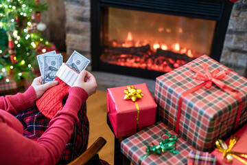 Naklejka na ściany i meble Woman putting american dollars in red christmas sock sitting near christmas tree and fireplace and packing gift box. concept.