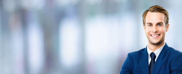 Portrait of happy confident businessman in blue suit and tie, standing against blurred modern office. Business success concept. Smiling man indoors. Copy space for some text or slogan.