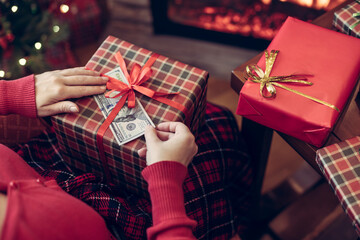 Woman sitting near christmas tree and fireplace and putting american dollars under red bow of huge gift box. Concept