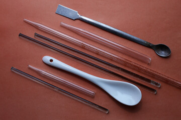 White ceramic spoon and other chemical tools: metal spatula, glass rods, tubes, and a measuring pipette. On a brown background.
