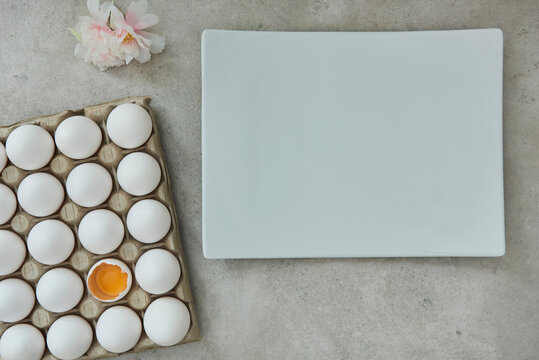 Egg Carton With A Cracked Egg, A Porcelain Plate And A Flower On A Gray Background