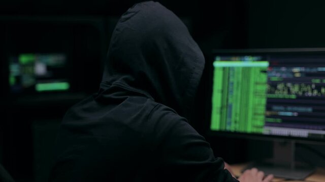 Back View Of Hooded Hacker Typing Code Surrounded By Multiple Monitors In Dark Office, Security Threat