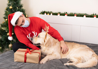 man wearing santa claus hat and medical mask hugs golden retriever in christmas atmosphere