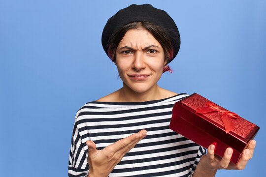 Studio Image Of Fashionable Young Female Frowning, Expressing Indignation, Holding Red Box, Gesturing Actively, Being Disappointed With Birthday Present. Displeased Girl Receiving Bad Gift