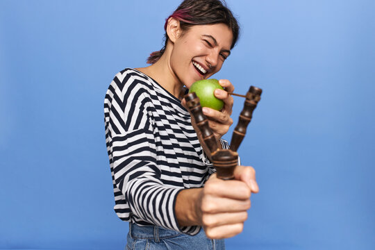 Portrait Of Playful Cute Teenage Girl In Sailor Shirt Having Fun Holding Slingshot, Pulling Back Elastic, Going To Launch Green Apple, Closing One Eyes, Aiming Right At Camera, Posing Isolated