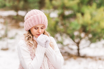 young woman warms her hands in the winter forest. She's wearing pink knitted clothes and white gloves.