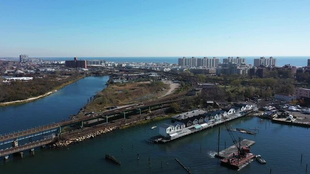 An Aerial Drone Shot Over Grassy Bay In A Neighborhood Of Queens, NY. The Camera Dolly In Over The Waters & Towards Elevated Train Tracks. There Are Houses & An Industrial Area In View On A Sunny Day.