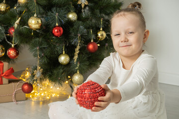 Cheerful cute child girl with Christmas boll having fun near Christmas tree wearing white dress.