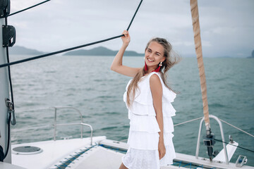 portrait of happy pretty girl with white dress, long red earrings and long curly blonde hair standing on yacht at summertime. looking at camera with toothy smile.
