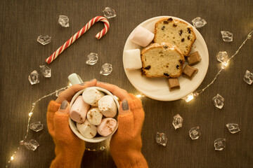 Girl's hands in knitted mittens hold a mug of hot drink with marshmallow on a table with plate with pieces of cake and chocolate, decorated with shining garland. Christmas atmosphere content