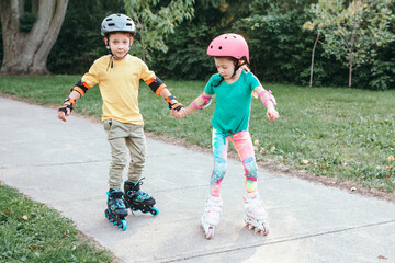 Happy Caucasian friends boy and girl in helmets riding on roller skates in park on summer day. Support and help from friend. Seasonal outdoors children activity sport. Kids individual summer sport.