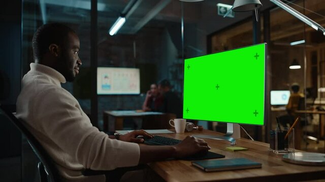 Handsome Black African American Specialist Working on Desktop Computer with Green Screen Mock Up Display in a Busy Creative Office. Male Manager is Wearing a Casual White Turtle Neck Sweater.