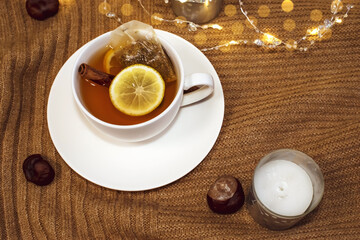 A white Cup of tea with lemon and cinnamon stands on a warm sweater near a candle and chestnuts. The concept of comfort and leisure