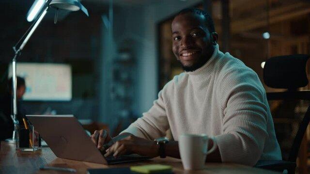 Handsome Black African American Man Having an Online Conversation on a Laptop Computer in Creative Office Environment. Happy Person of Color in Warm White Sweater is Looking at Camera and Smiling.