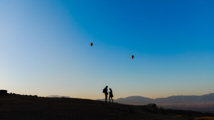 Naklejka na ściany i meble Silhouette of a man and a woman on a mountainside at early morning sunrise. Balloons fly over them in the sky. A romantic painting against the backdrop of a beautiful landscape. High quality photo