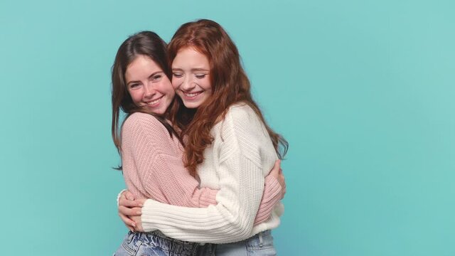 Two cute happy young brunette redhead women friends 20s years old in casual clothes pastel pink white sweaters hugging embracing each other isolated on blue background studio. People lifestyle concept