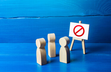 A group of people figurines looking at an easel with a red prohibitory symbol NO. Expression of...
