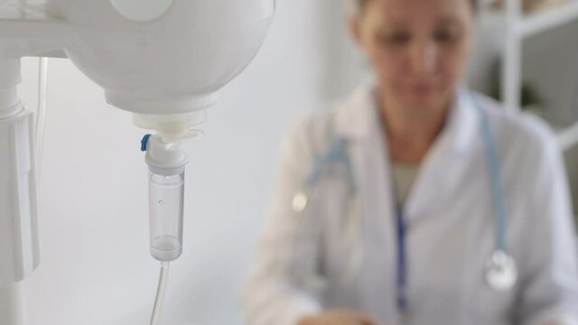 Close up of IV dripper with saline and elderly female doctor sitting on background