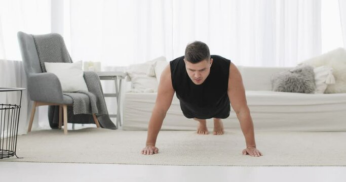 Sporty athletic male exercising on his hard workout on quarantine. Intense training in modern light living room. Young man is doing push-ups exercises at home keeping himself during lockdown.
