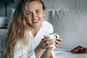 Beautiful blonde woman in white coat with white smile having breakfast in a hotel room. Woman is...