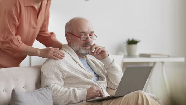 Medium shot of senior Caucasian man sitting on couch in living room, using laptop. Then his wife coming, standing behind him and starting hugging him and kissing on cheek