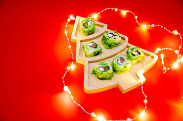 The idea of ​​a festive sushi bar menu. creative Christmas tree made of green rolls lying on a wooden plate in the shape of a Christmas tree on a red background. glowing lights around the tree