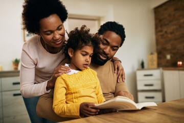 Small black girl reading a book with her parents at home.