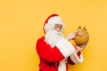 Funny and cute Santa Claus is holding a stuffed reindeer toy in his hand, touching a nose of the toy. Amusing Santa is playing with a teddy on a yellow background.