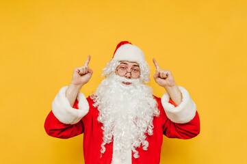 Fototapeta na wymiar Cheerful and positive Santa Claus is pointing up on a copy space. Santa on a yellow background is raising his hands up, advertising and bringing attention to a blank space.