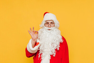 Fototapeta na wymiar Portrait of a bearded Santa Claus showing an ok gesture and looking at the camera. Santa is expressing an ok symbol with his hand, standing on a yellow background.