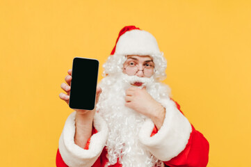 Santa Claus is bringing up attention by showing a smartphone, holding a device in his hand, looking interested. Focus on a mobile phone.