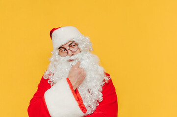Close up portrait of Santa Claus on a yellow background, touching his beard, pondering over Christmas holidays, making a thinking gesture, looking interested.