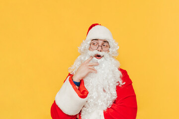 Thoughtful Santa Claus is standing at the studio on the yellow background, touching his beard with a thinking gesture, looking aside with his mouth open.
