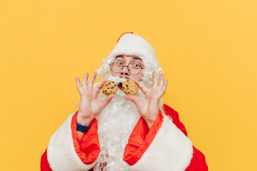Funny cheerful Santa Claus is holding chocolate cookies in his hands, looking at the camera, feeling surprised, eyes and mouth wide open. Man in Santa costume is standing in front of the yellow wall.