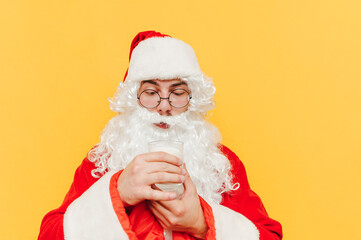 Closeup portrait of Santa Claus holding a glass of milk, looking at milk, feeling delighted and curious, standing on a yellow background.
