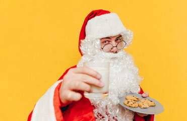 Closeup portrait of Santa Claus on a yellow background holding a glass of milk and a plate with chocolate cookies, offering milk to the viewer.