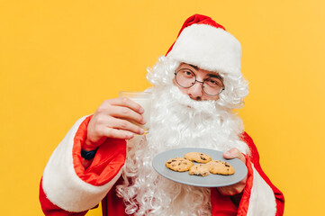 Old Santa Claus is holding a glass of milk and a plate with chocolate cookies, offering those goodies to the viewer. Close up portrait, isolated on a yellow background.