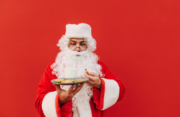 Concentrated Santa Claus is standing in front of a red wall, holding a plate with biscuits and a glass of milk, taking one cookie and going to eat it.