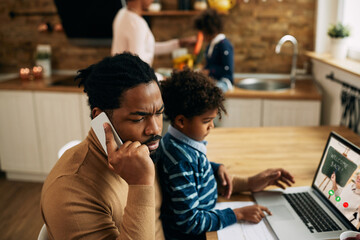 African American man talking on the phone while his son is using laptop and following online class.
