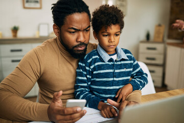 African American stay at home father using phone while working on laptop.